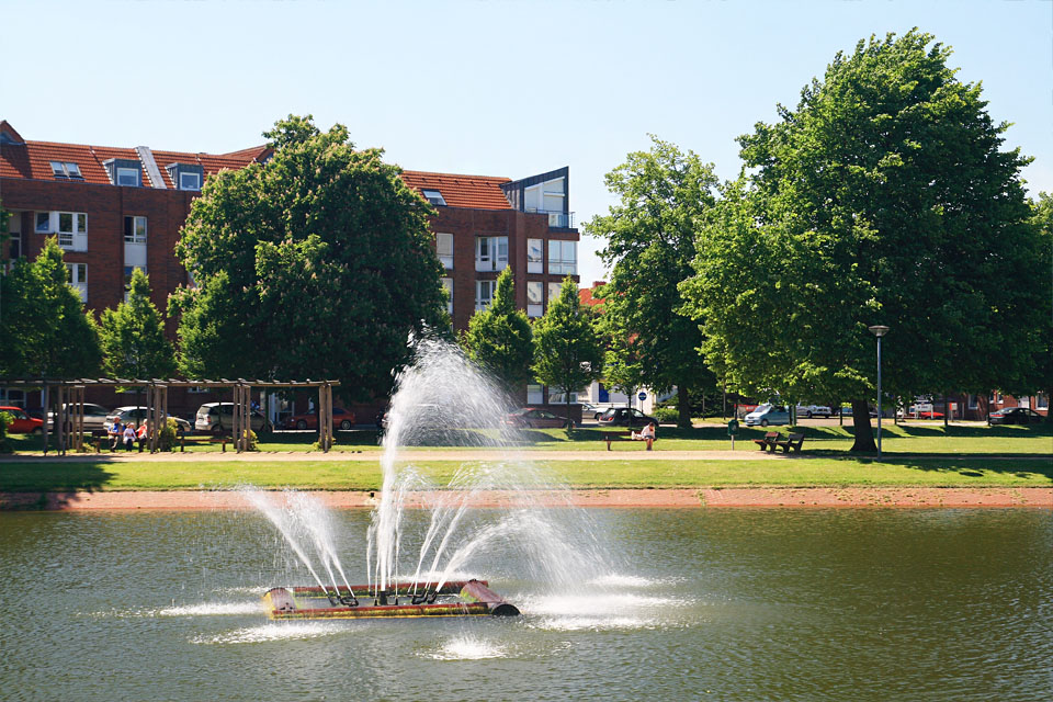Ein Mehrfamilienhaus und davor ein See mit einem Springbrunnen.